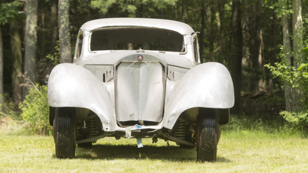 Ein Oldtimer mit silberner Karosserie steht auf einer Wiese vor einem Wald.
