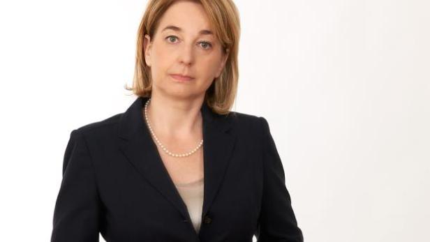 Eine Frau mit blonden Haaren und einer Perlenkette trägt ein dunkles Jackett.