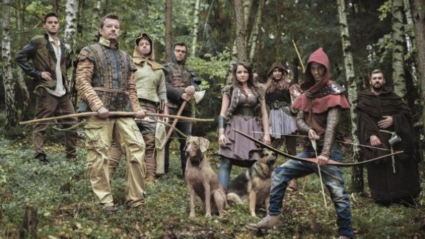 Eine Gruppe von Personen in mittelalterlichen Kostümen posiert mit Bögen und Hunden in einem Wald.