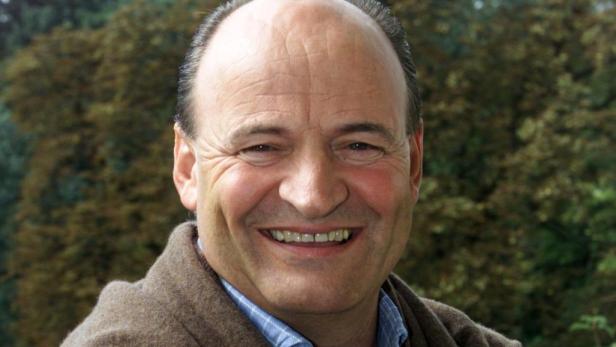 Ein lächelnder Mann mit brauner Jacke vor einem grünen Hintergrund.