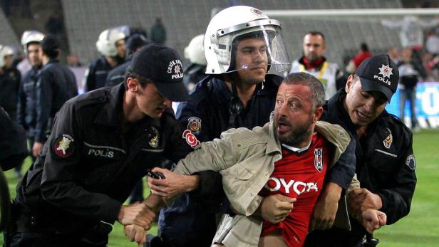 Polizisten führen einen Mann in einem roten Trikot vom Fußballfeld ab.