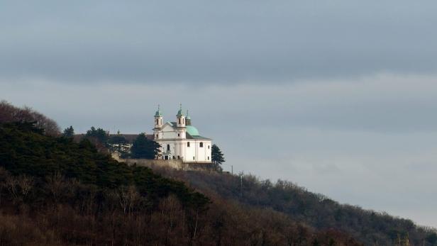 Eine weiße Kirche mit grünen Kuppeln thront auf einem bewaldeten Hügel.