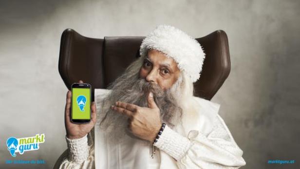Ein Mann mit Bart und weißem Gewand präsentiert die „Marktguru“-App auf einem Smartphone.