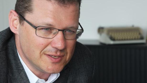 Ein Mann mit Brille und grauem Sakko vor einem Schreibtisch mit einer Schreibmaschine.