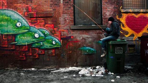 Ein Mann sitzt auf einer Mülltonne und angelt vor einer mit Graffiti bemalten Wand.