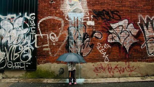 Ein Kind mit Regenschirm steht vor einer mit Graffiti bedeckten Ziegelmauer.