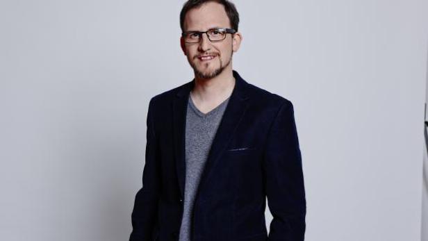 Ein Mann mit Brille und Bart trägt ein blaues Sakko und ein graues T-Shirt.