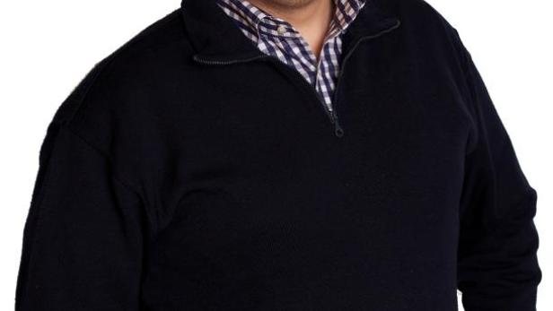 Ein lächelnder Mann mit Brille, blauem Pullover und Jeans posiert vor einem weißen Hintergrund.