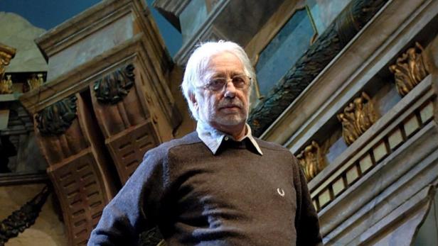Ein Mann mit Brille und grauem Haar steht vor einer Kulisse mit antiken Architekturelementen.