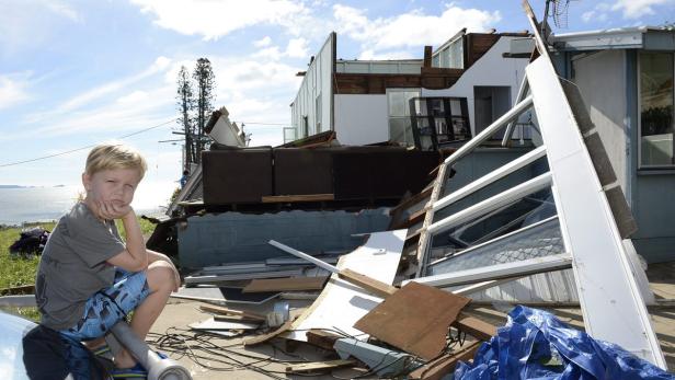 Ein Junge sitzt vor einem durch einen Sturm beschädigten Haus.