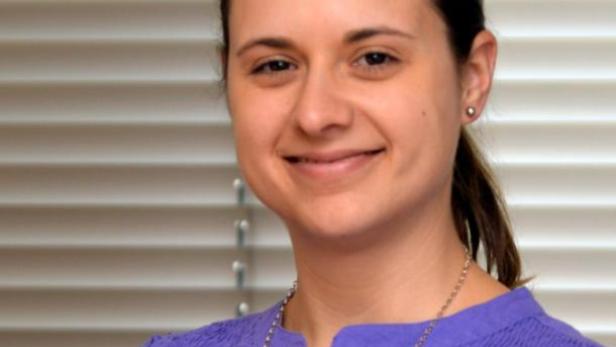 Eine lächelnde Frau mit braunen Haaren und einem lila Oberteil steht vor einem Fenster mit Jalousien.