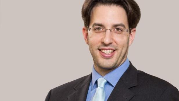 Ein lächelnder Mann mit Brille, Anzug und hellblauem Hemd vor einem grauen Hintergrund.