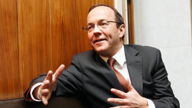 Ein Mann mit Brille und Anzug gestikuliert vor einer Holzwand.