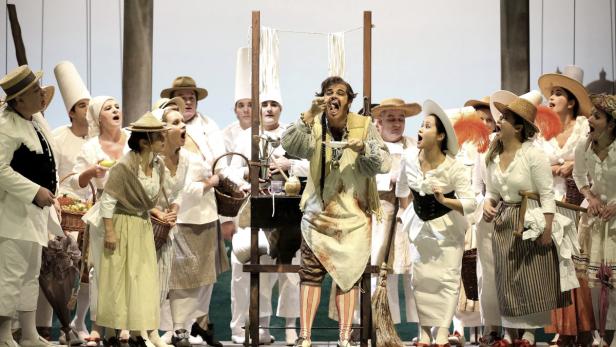 Eine Opernszene mit einem Mann, der Nudeln isst, umgeben von einem Chor.