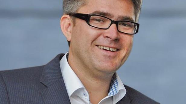 Ein lächelnder Mann mit Brille und grau meliertem Haar trägt einen Anzug.