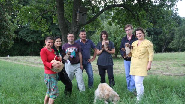 Eine Gruppe von Menschen steht mit Stofftieren und einem Hund auf einer Wiese unter einem Baum mit einem Vogelhaus.
