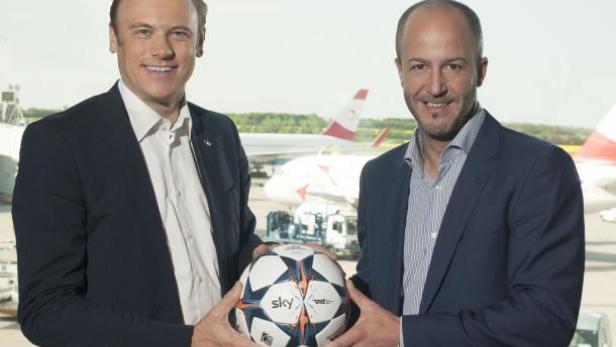 Zwei Männer halten einen Fußball mit dem Sky-Logo vor einem Flughafen.