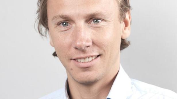Ein lächelnder Mann mit blonden Haaren und blauem Hemd vor einem grauen Hintergrund.
