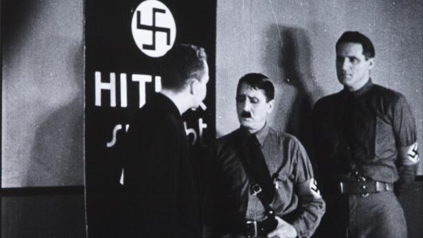 Eine Schwarzweißaufnahme von Adolf Hitler im Gespräch mit zwei Männern vor einem Plakat.
