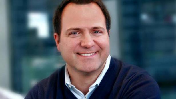 Ein lächelnder Mann mit dunklen Haaren und einem blauen Pullover.