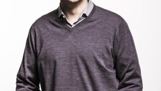 Ein lächelnder Mann mit Brille, Pullover und Jeans posiert vor einem weißen Hintergrund.