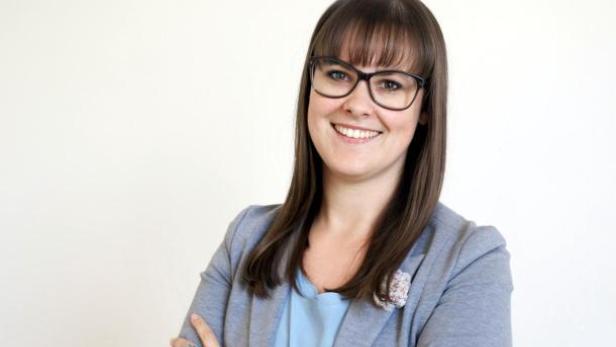 Eine lächelnde Frau mit Brille und braunen Haaren vor einem weißen Hintergrund.