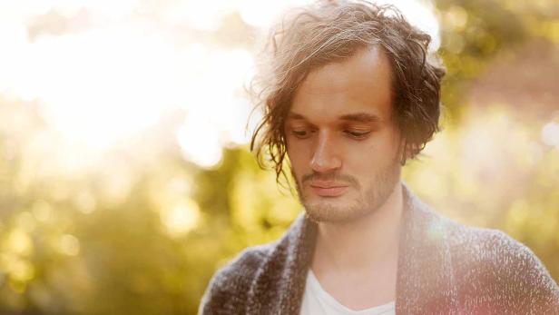 Ein Mann mit lockigem Haar und Bart trägt eine Strickjacke.