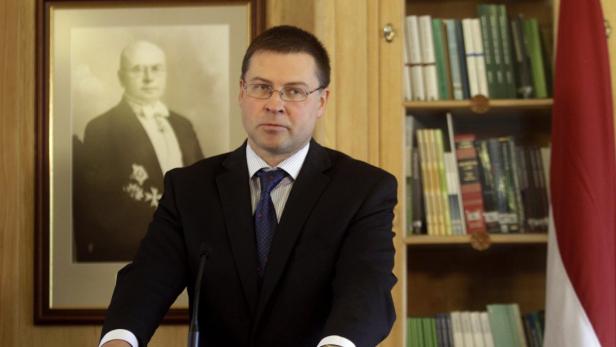 Ein Mann in Anzug und Krawatte steht vor einem Bücherregal und einem Porträt.