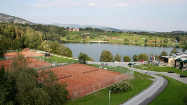 Blick auf Tennisplätze und einen See in einer grünen Landschaft.