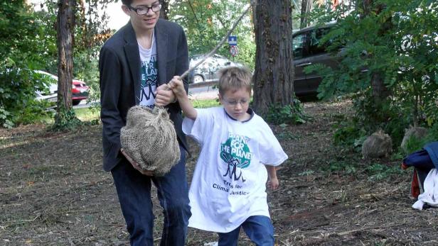 Zwei Kinder tragen einen Sack und einen Stock durch einen Wald, möglicherweise zum Pflanzen von Bäumen.