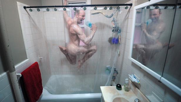 Ein Mann springt nackt unter der Dusche und singt in eine Shampooflasche.