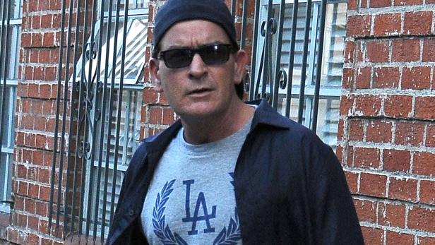 Charlie Sheen mit Sonnenbrille und Baseballkappe vor einer Backsteinmauer.