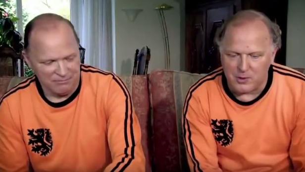 Zwei Männer, vermutlich Zwillinge, sitzen nebeneinander und tragen orangefarbene Trikots mit einem Löwenemblem.