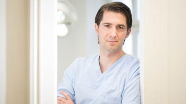 Ein Mann in hellblauer Arztkleidung lehnt an einer Wand.