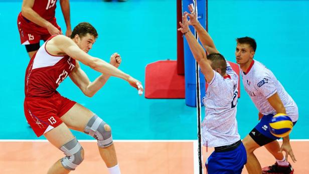 Ein Volleyballspieler schlägt den Ball über das Netz, während zwei Gegenspieler blocken wollen.