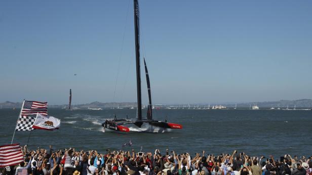 Ein Katamaran der Oracle Team USA fährt vor einer jubelnden Menschenmenge und Flaggen.
