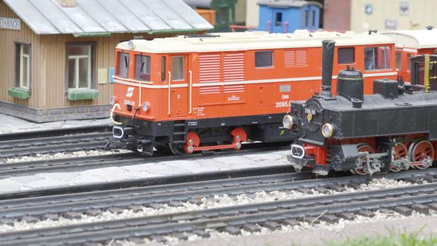 Eine orangefarbene Elektrolokomotive und eine schwarze Dampflokomotive auf einem Modellbahngleis.