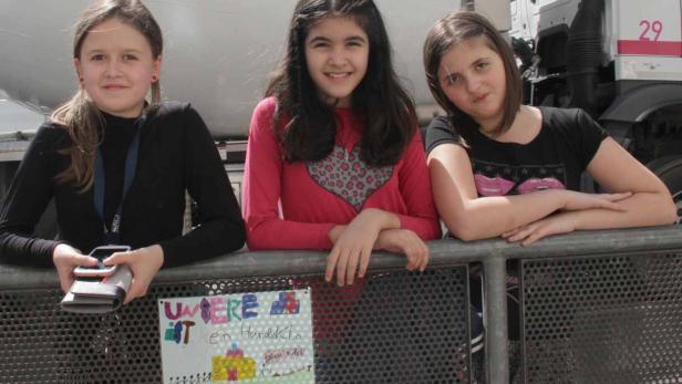 Drei Mädchen stehen lächelnd an einer Absperrung vor einem LKW.