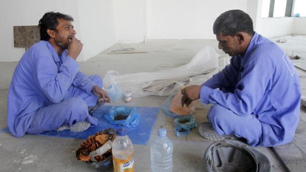 Zwei Bauarbeiter in blauen Overalls machen eine Mittagspause auf dem Boden.