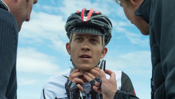 Ein Radrennfahrer mit Helm wird von zwei Personen unterstützt.