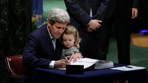 John Kerry unterschreibt ein Dokument, während er ein kleines Mädchen im Arm hält.