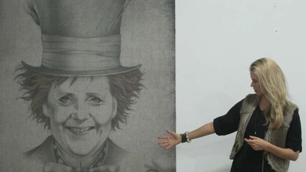 Eine Frau deutet auf ein Porträt von Angela Merkel mit Zylinder und Fliege.