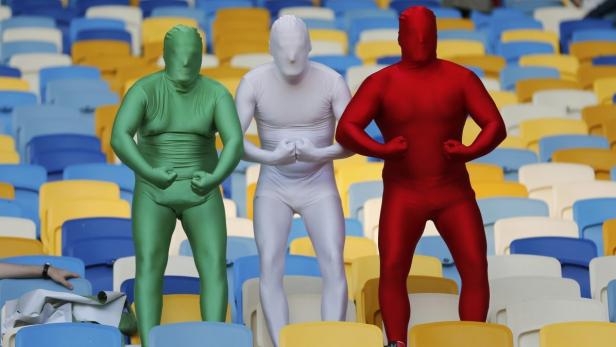 Drei Personen in Ganzkörperanzügen in den Farben Grün, Weiß und Rot posieren auf einer Tribüne.