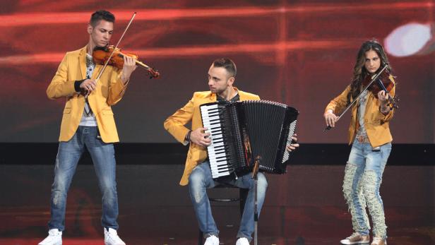 Drei Musiker in gelben Jacken spielen Geige und Akkordeon auf einer Bühne.