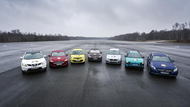 Sieben Autos unterschiedlicher Marken stehen auf einem grauen Asphaltplatz nebeneinander.