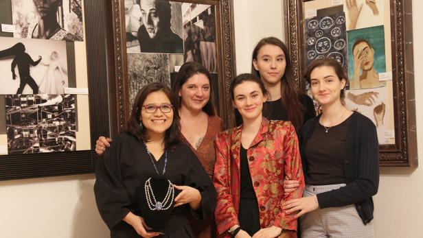 Fünf Frauen posieren vor einer Wand mit gerahmten Kunstwerken.