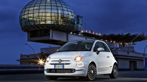 Ein weißer Fiat 500 vor einem Gebäude mit einer gläsernen Kuppel bei Nacht.