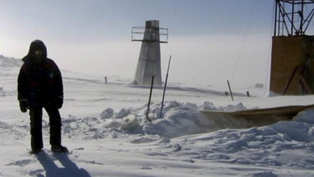 Eine Person steht in einer verschneiten Landschaft mit Forschungsstationen im Hintergrund.