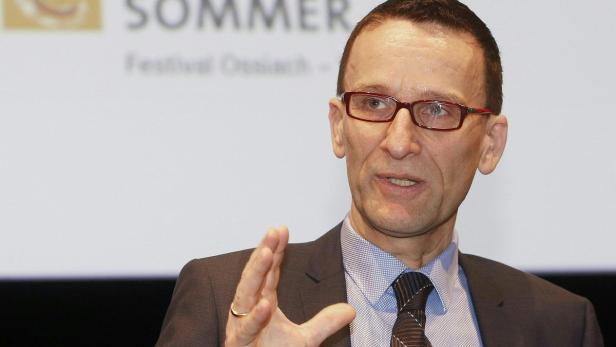 Ein Mann mit Brille gestikuliert vor einem Banner des Festivals Ossiach.