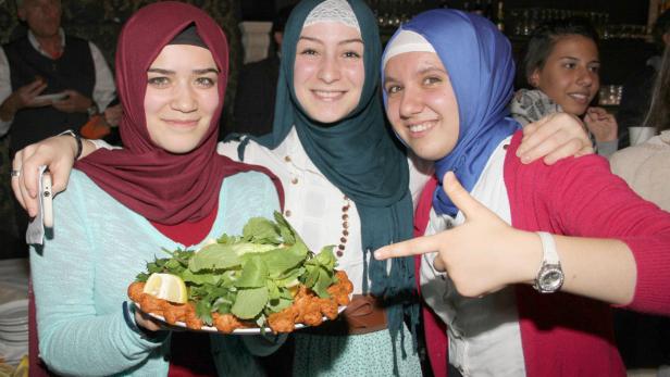 Drei lächelnde junge Frauen mit Kopftüchern präsentieren eine Platte mit türkischen Köfte und Salat.